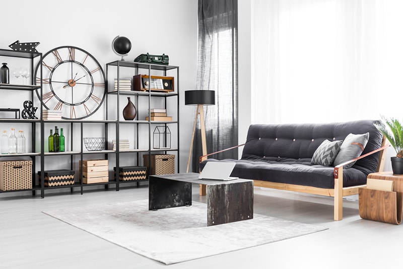 Laptop on table and wooden sofa in bright living room interior with industrial clock and bottles on a shelf